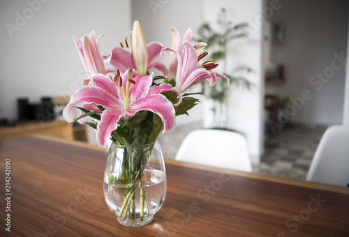 Bouquet pink lily flowers in glass vase on wood table in room.