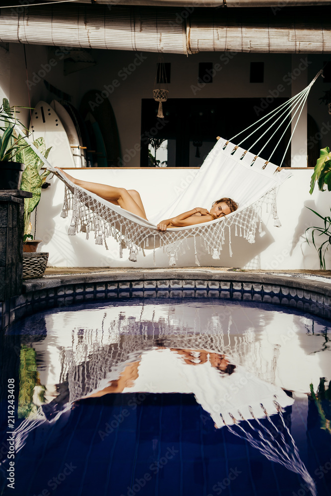 Fototapeta premium Young beautiful sporty lady rest and swinging in white hammock and reflect in the blue pool. Tropical private house. Cheerful woman enjoy her vacation after hard office day to day work.