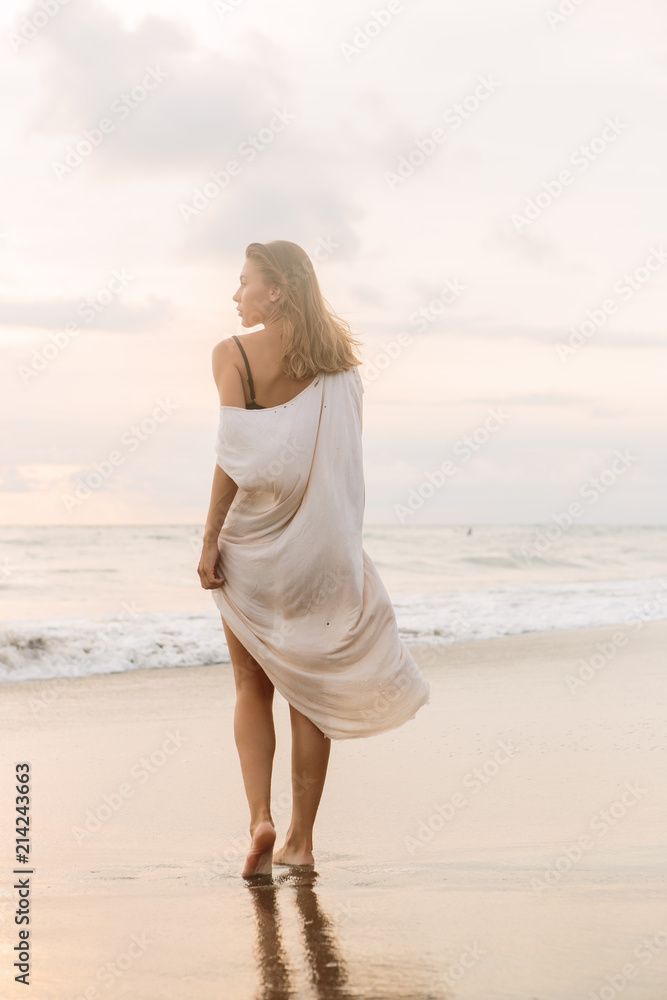 Hipster trendy girl in casual summer dress walking barefoot by the waterline and look to the waves. Sporty lady on sea sand beach sunset or ocean sunrise. Travel, active, yoga lifestyle concept. Hipster trendy girl in casual summer dress walking barefoot by the waterline and look to the waves. Sporty lady on sea sand beach sunset or ocean sunrise. Travel, active, yoga lifestyle concept.