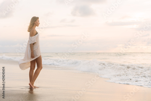 Hipster trendy woman in casual summer dress walking barefoot by the waterline and look to the waves. Sporty lady on sea sand beach sunset or ocean sunrise. Travel, active, yoga lifestyle concept.