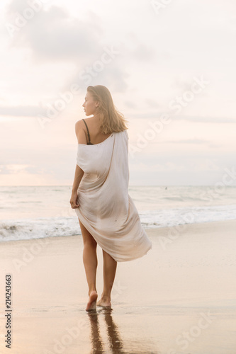 Hipster trendy girl in casual summer dress walking barefoot by the waterline and look to the waves. Sporty lady on sea sand beach sunset or ocean sunrise. Travel  active  yoga lifestyle concept.