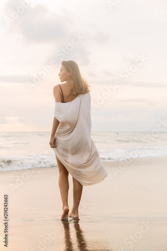 Hipster trendy girl in casual summer dress walking barefoot by the waterline and look to the waves. Sporty lady on sea sand beach sunset or ocean sunrise. Travel, active, yoga lifestyle concept.