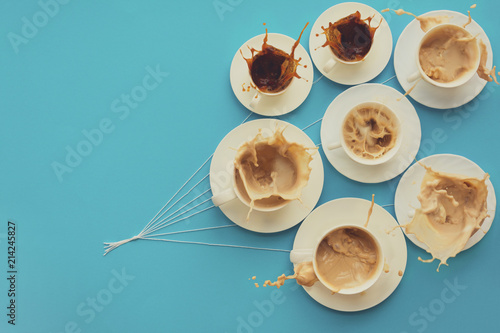Hand holding coffee cups with milk and without in shape of balloons on blue paper background. Toned. Weather or good morning concept.