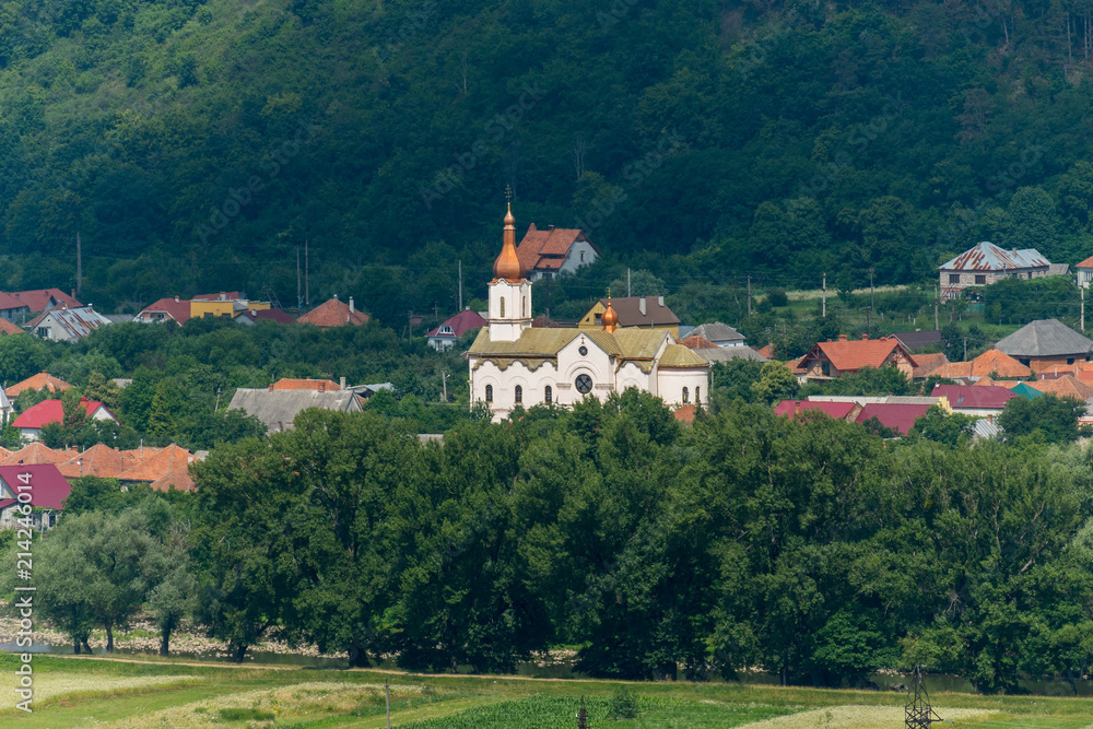 Church with white walls and bulbs of domes with nearby village houses among the trees in a picturesque area on the background of a mountain slope.