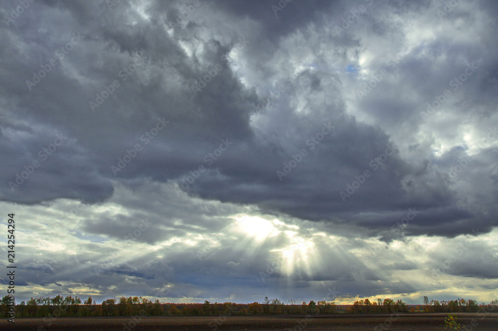 Cloudy sky with thick dense clouds that tightened the whole sky and thin rays of the sun trying to break through them on the field with the trees below. Cloudy sky with thick dense clouds that tightened the whole sky and thin rays of the sun trying to break through them on the field with the trees below.