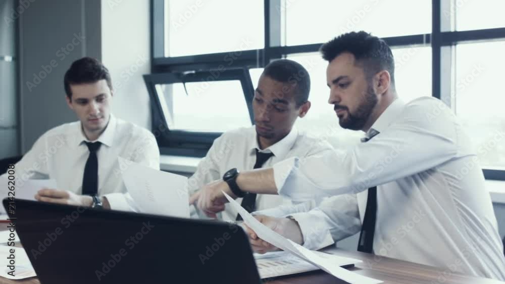 Young team of three businessmen, two white and one african national, sit in shirts at table and discuss project, papers with charts. Work of business team. Brainstorm. Panoramic windows in office. Young team of three businessmen, two white and one african national, sit in shirts at table and discuss project, papers with charts. Work of business team. Brainstorm. Panoramic windows in office.