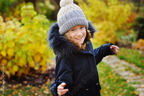 autumn portrait of happy kid girl walking in the garden in warm parka coat and knitted hat