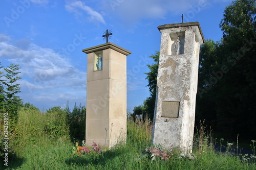 Dwie murowane kapliczki przydrożne, jedna zabytkowa, stara, zniszczona, pochylona, obok niej nowa, stoi prosto, w tle zieleń, błękitne niebo