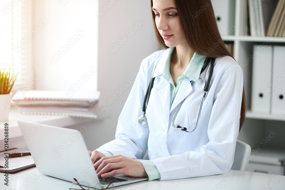 Young female doctor typing on laptop computer while sitting at the table near the window in hospital office Young female doctor typing on laptop computer while sitting at the table near the window in hospital office