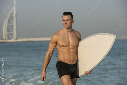Handsome muscular athletic white caucasian male showing six pack abs on walking out of the water on the beach with surf board with crashing waves in Dubai