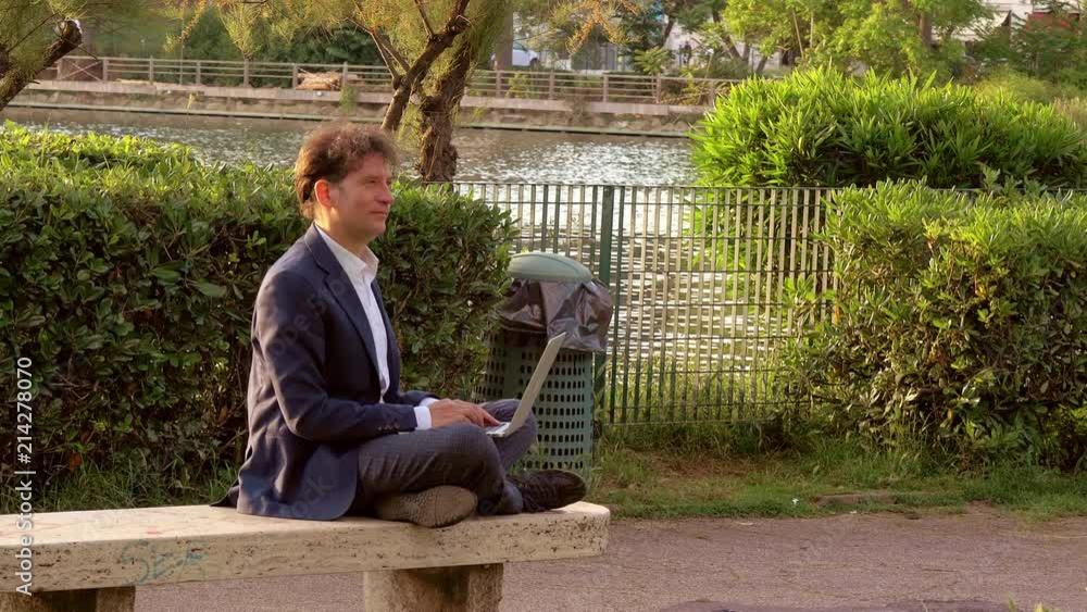 Man in park in front of lake working with pc at sunset slow motion breathing Man in park in front of lake working with pc at sunset slow motion breathing