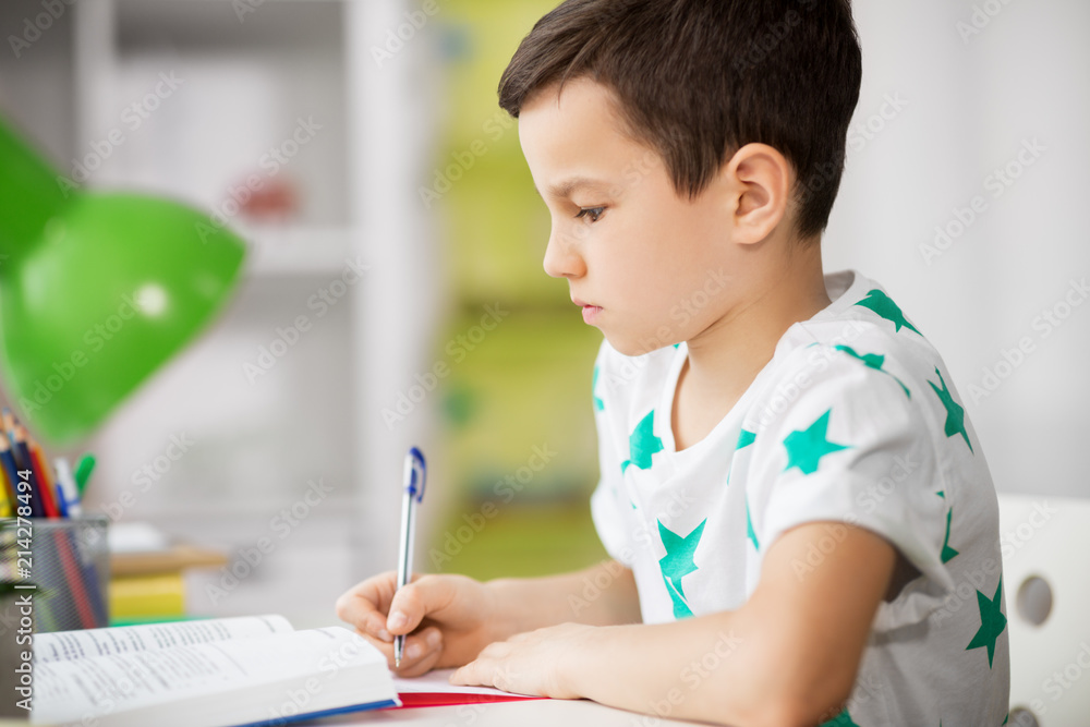 education, childhood and school concept - close up of boy with textbook writing to notebook at home