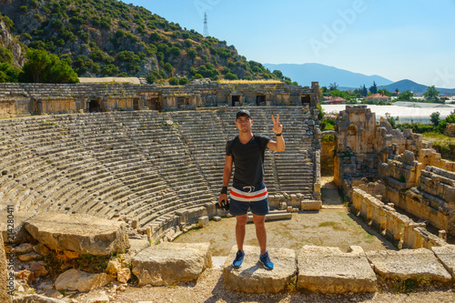 A young man in shorts, a gray t-shirt and a black cap stands against the background of an ancient amphitheater with a camera in his hand and shows a symbol of peace