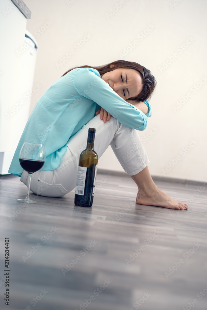 Sad woman sitting on the floor hugging her knees with a bottle of red ...