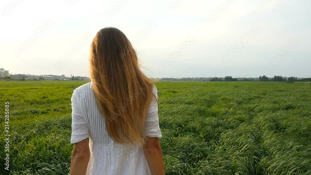 Beautiful woman in white dress walks on the green field. Wind blowing hair. Grassy summerfield on sunset. Slow motion Beautiful woman in white dress walks on the green field. Wind blowing hair. Grassy summerfield on sunset. Slow motion