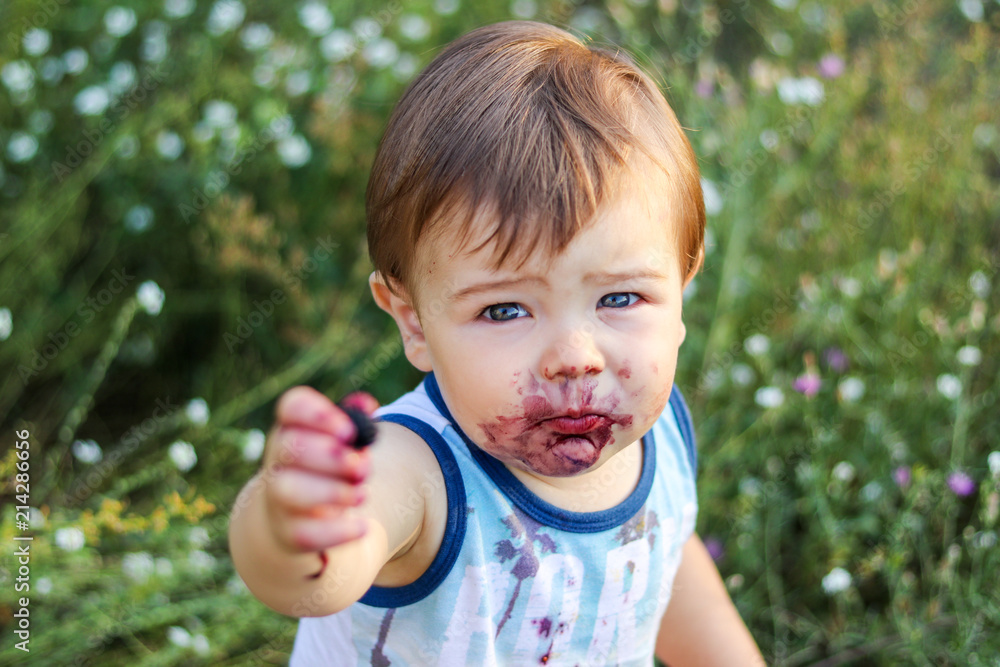 Adorable little baby boy with face smeared around his mouth with mulberry juice offering a berry looking at camera outdoors. Selective focus. Happy childhood concept Adorable little baby boy with face smeared around his mouth with mulberry juice offering a berry looking at camera outdoors. Selective focus. Happy childhood concept