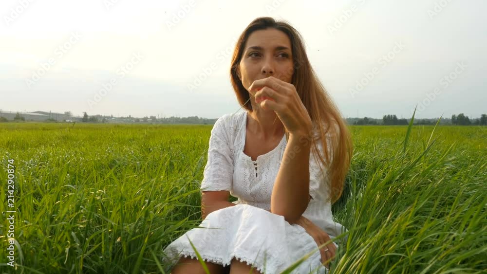 Portrait happy young woman on a summer walk in field. Cute girl sitting on the green grass and dreaming. Beautiful young woman enjoying nature in summer evening. Grassy summerfield Portrait happy young woman on a summer walk in field. Cute girl sitting on the green grass and dreaming. Beautiful young woman enjoying nature in summer evening. Grassy summerfield