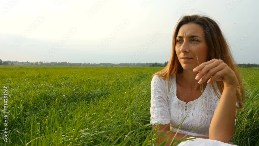 Portrait happy young woman on a summer walk in field. Cute girl sitting on the green grass and dreaming. Beautiful young woman enjoying nature in summer evening. Grassy summerfield Portrait happy young woman on a summer walk in field. Cute girl sitting on the green grass and dreaming. Beautiful young woman enjoying nature in summer evening. Grassy summerfield