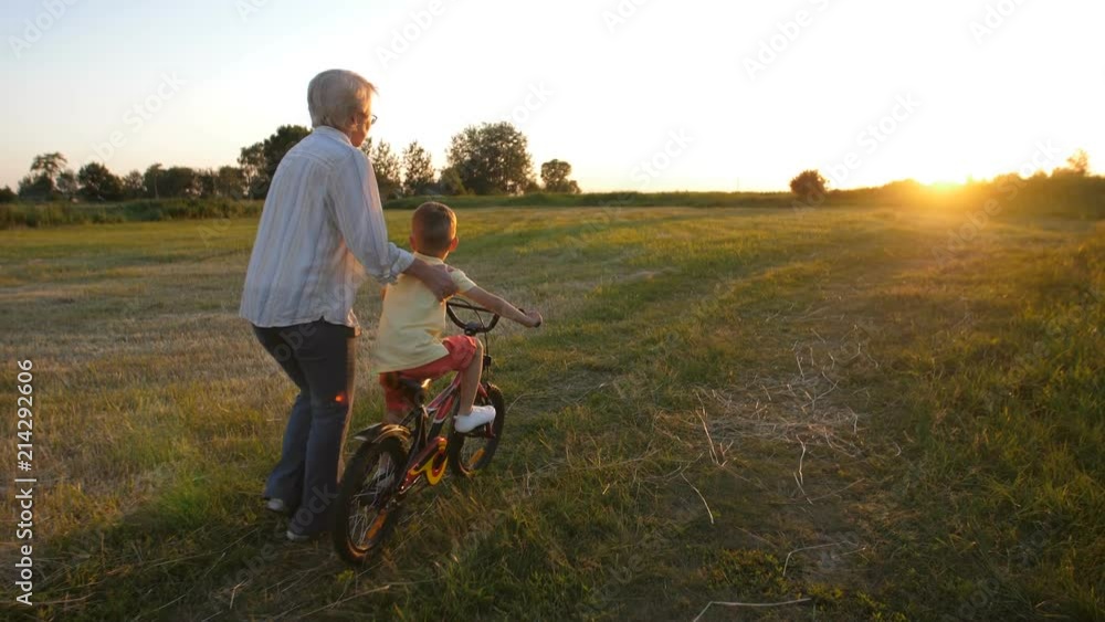 Back view of toddler boy learning to ride a bicycle with grandmother's help. Cute blond grandchild pressing bike pedals while granny holding and keeping him safe. Loving grandmother helping grandson Back view of toddler boy learning to ride a bicycle with grandmother's help. Cute blond grandchild pressing bike pedals while granny holding and keeping him safe. Loving grandmother helping grandson