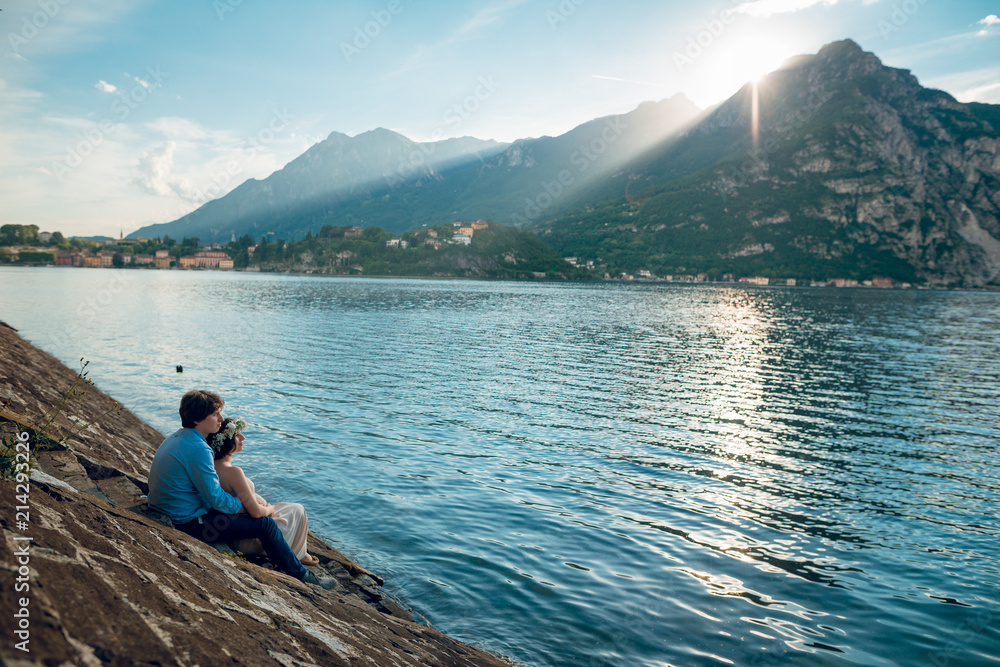 Young couple of pregnant woman and man sitting on the stairs on the shore of Lake Como with wonderful mountain view on sunny summer day