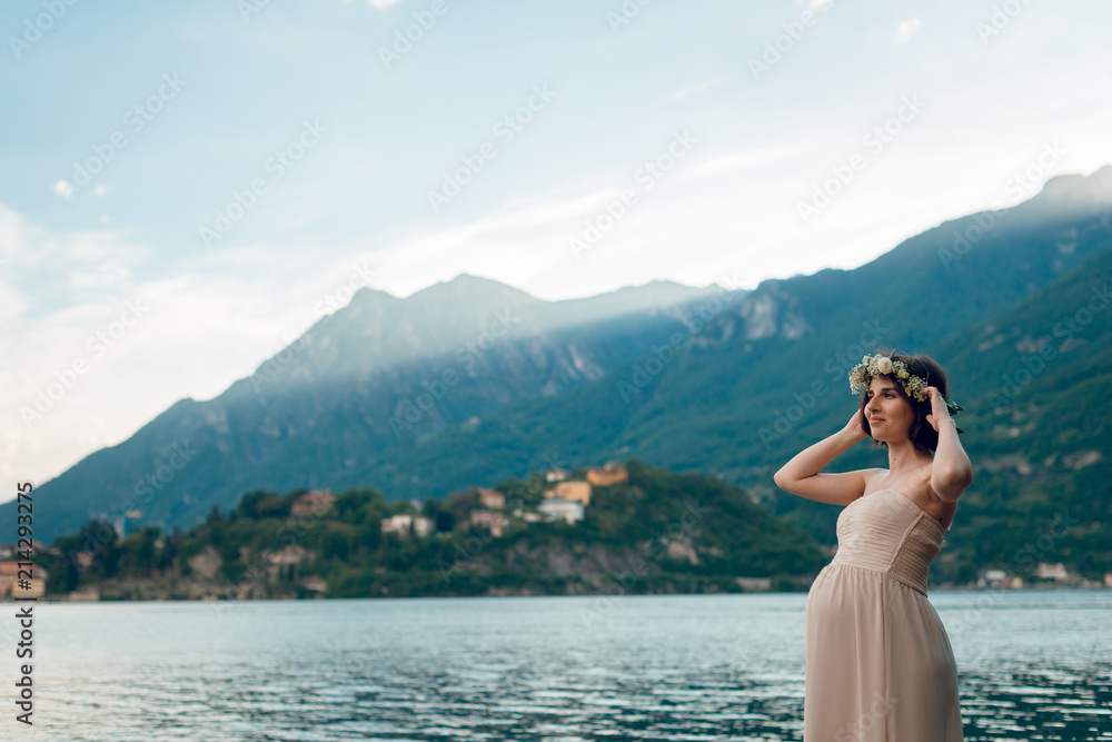 Beautiful pregnant woman in creme dress and flower wreath standing on the shore with scenic view of the lake Como and green mountains on background