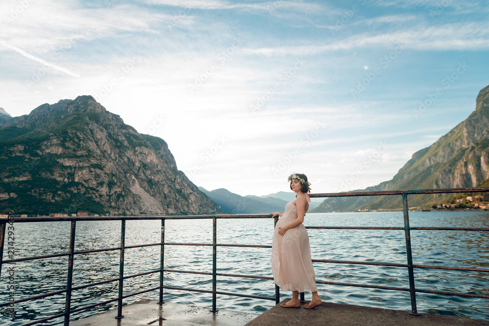 Young tender pregnant woman in nice dress standing on the pier near the Como lake. Scenic view beautiful mountains and lake. Summer day