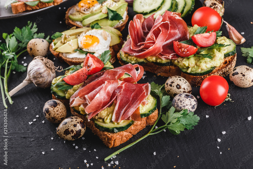 Various of sandwiches and bruschetta with prosciutto, fried quail egg, avocado, cucumber, tomatoes, on black stone background.