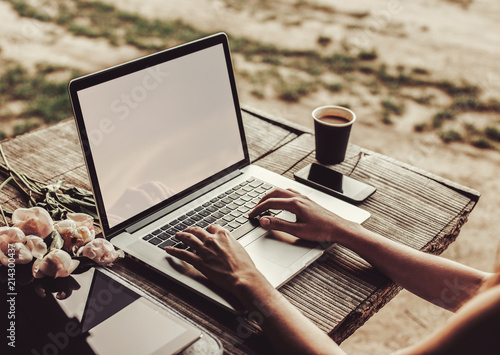 Young woman using and typing laptop computer at rough wooden table with coffee cup, strawberries, bouquet of peonies flowers photo