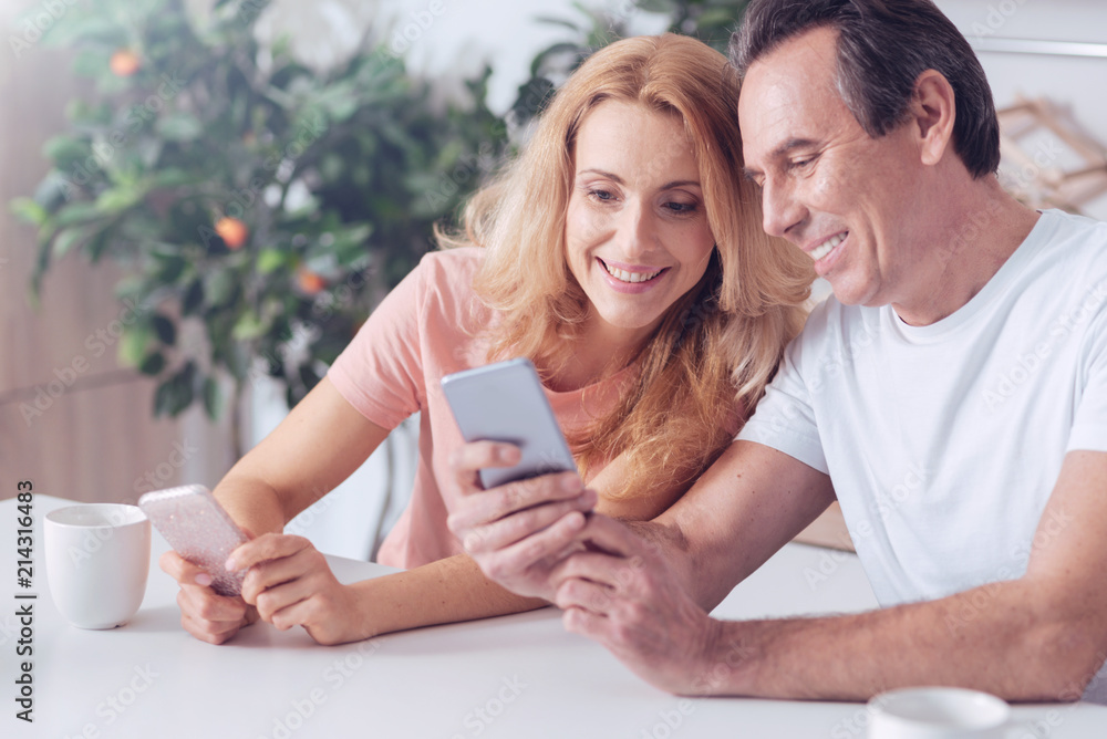 Happy marriage. Delighted positive cheerful man sitting together with his wife and holding his smartphone while showing her an interesting picture Happy marriage. Delighted positive cheerful man sitting together with his wife and holding his smartphone while showing her an interesting picture