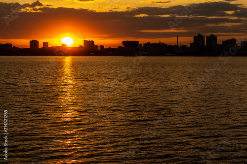 sunset over the river in the city with silhouettes of buildings and the reflection of the sun in the water © Иван Ульяновский