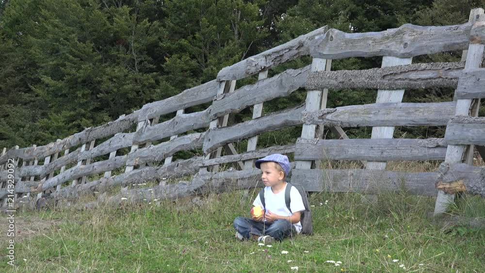 Lovely child with a backpack eating apple, resting on green grass near wooden fence Lovely child with a backpack eating apple, resting on green grass near wooden fence