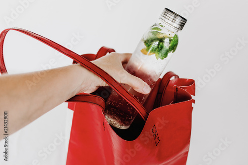 Transparent bottle with water infused with lemon and peppermint leaves to take with on hot summer days. Body parts, hand putting the bottle into red tote bag. White background, isolated, copy space.