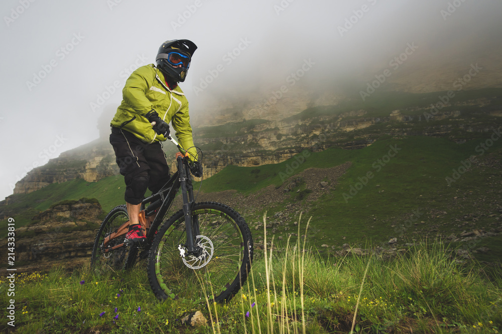 An adult mtb cyclist on a mountain bike at the foot of a cliff surrounded by green grass. Low clouds. North Caucasus. Russia An adult mtb cyclist on a mountain bike at the foot of a cliff surrounded by green grass. Low clouds. North Caucasus. Russia