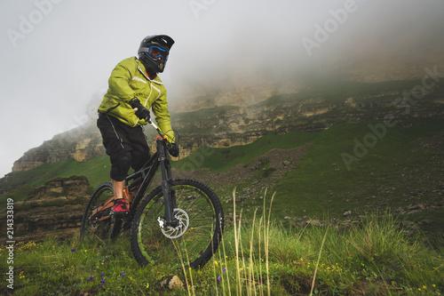 An adult mtb cyclist on a mountain bike at the foot of a cliff surrounded by green grass. Low clouds. North Caucasus. Russia