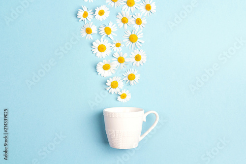 White cup and chamomile flowers on a blue background. Chamomiles come out of the cup like steam. Chamomile tea concept
