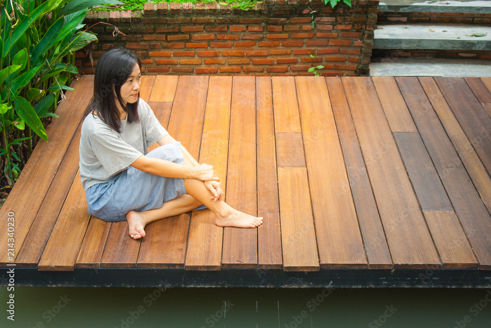 Lonely Asian woman sitting relax on wooden terrace or porch near pond in the garden.