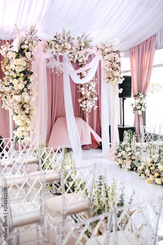 Fototapeta premium The wedding ceremony area is decorated with white and peach cloth, crystal chandelier, transparent chairs for guests and beautiful floral arrangements of roses