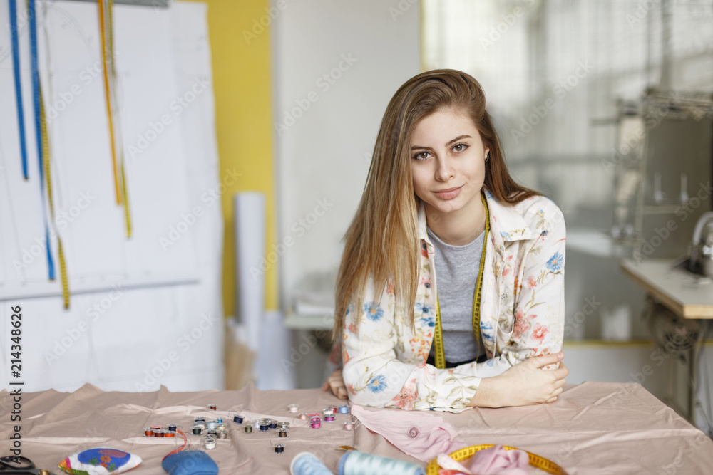 Pretty young seamstress in shirt leaning on table and dreamily looking in camera in modern sewing workshop
