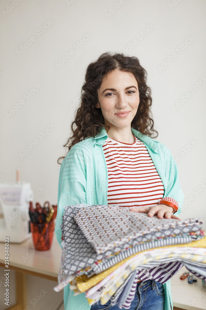 Beautiful smiling seamstress joyfully looking in camera while holding patterns fabrics in hands in sewing workshop Beautiful smiling seamstress joyfully looking in camera while holding patterns fabrics in hands in sewing workshop