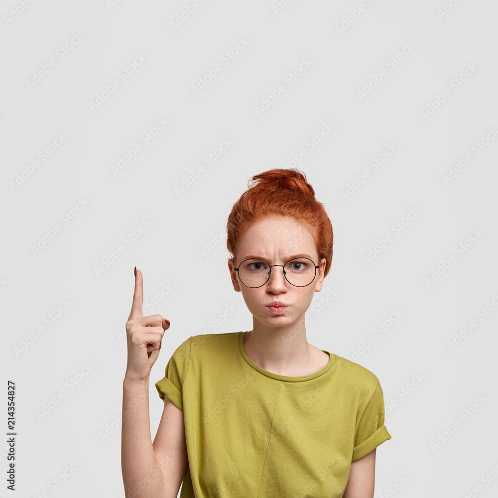 Angry female with ginger hair combed in bun, points upwards with fore finger, shows blank space for your advertisement or promotional text, wears spectacles and green t shirt, has sullen expression Angry female with ginger hair combed in bun, points upwards with fore finger, shows blank space for your advertisement or promotional text, wears spectacles and green t shirt, has sullen expression