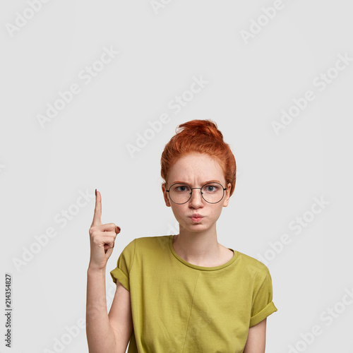 Angry female with ginger hair combed in bun, points upwards with fore finger, shows blank space for your advertisement or promotional text, wears spectacles and green t shirt, has sullen expression