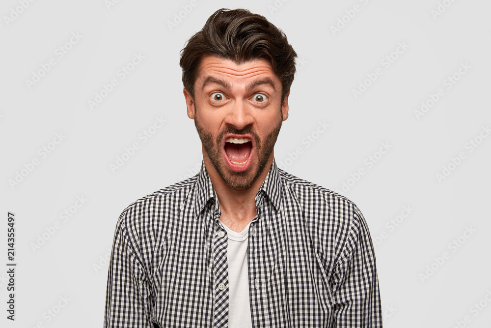 Obraz premium Aggressive furious bearded male has annoyed facial expression, raises eyebrows, shouts at someone during quarrel, argues in irritation, has dark hair, isolated over white background, dressed in shirt