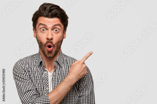 Fotografie Fashionable young male with stubble, has surprised expression, points aside at blank space for your advertisement or promotional text, expresses wonder and shock