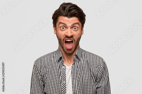 Aggressive furious bearded male has annoyed facial expression, raises eyebrows, shouts at someone during quarrel, argues in irritation, has dark hair, isolated over white background, dressed in shirt © Wayhome Studio