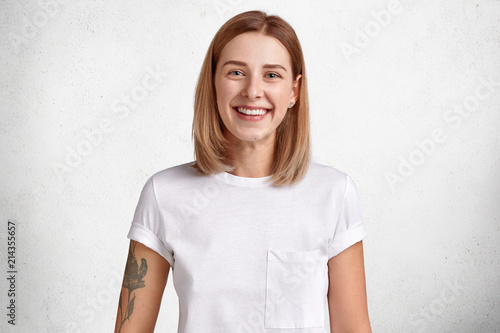 Waist up portrait of cheerful young female with short hair, bright smile, has tattoo on arm, rejoices positive news, stands against white concrete wall. People, emotions and happiness concept.