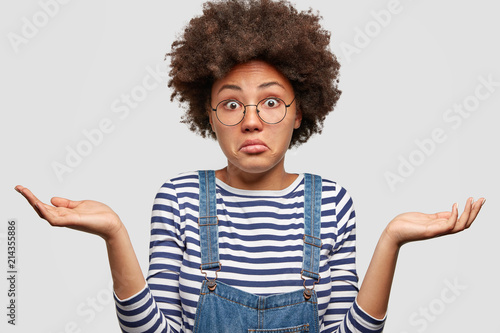 Quadro em tela Clueless African American female with curly bushy hair, shrugs shoulders, looks doubtfully as faces dilemma, wears striped sweater and jean overalls, stands against white background