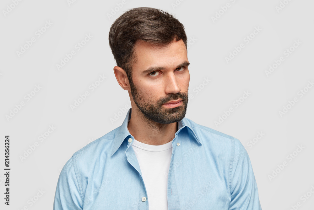 Confident unshaven guy with dark bristle and hair, listens attentively boss, stands against white background wears blue shirt. Serious male entrepreneur decides on new plan. Facial expressions concept Confident unshaven guy with dark bristle and hair, listens attentively boss, stands against white background wears blue shirt. Serious male entrepreneur decides on new plan. Facial expressions concept