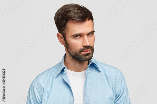 Confident unshaven guy with dark bristle and hair, listens attentively boss, stands against white background wears blue shirt. Serious male entrepreneur decides on new plan. Facial expressions concept