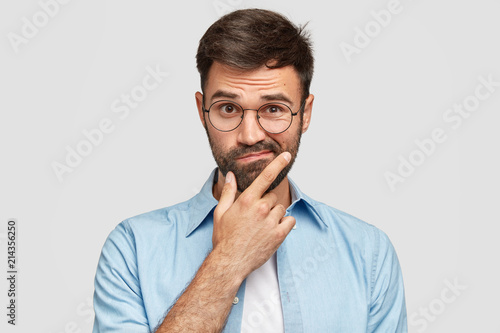 Fototapeta Image of hesitant unshaven European male with thick beard, holds chin, purses lips with clueless expressions, doubts what to eat delicious for supper, isolated over white background