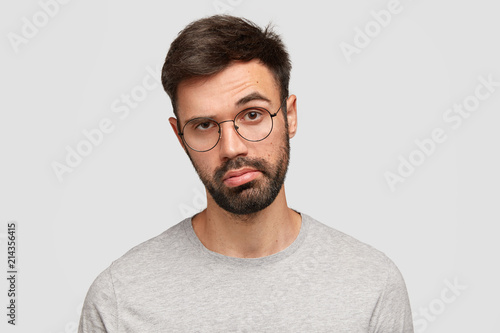 Indoor shot of good looking unshaven young male has bored hesitant expression, raises eyebrow in bewilderment, purses lips, makes decision, feels sick and tired of bachelorhood and being alone photo